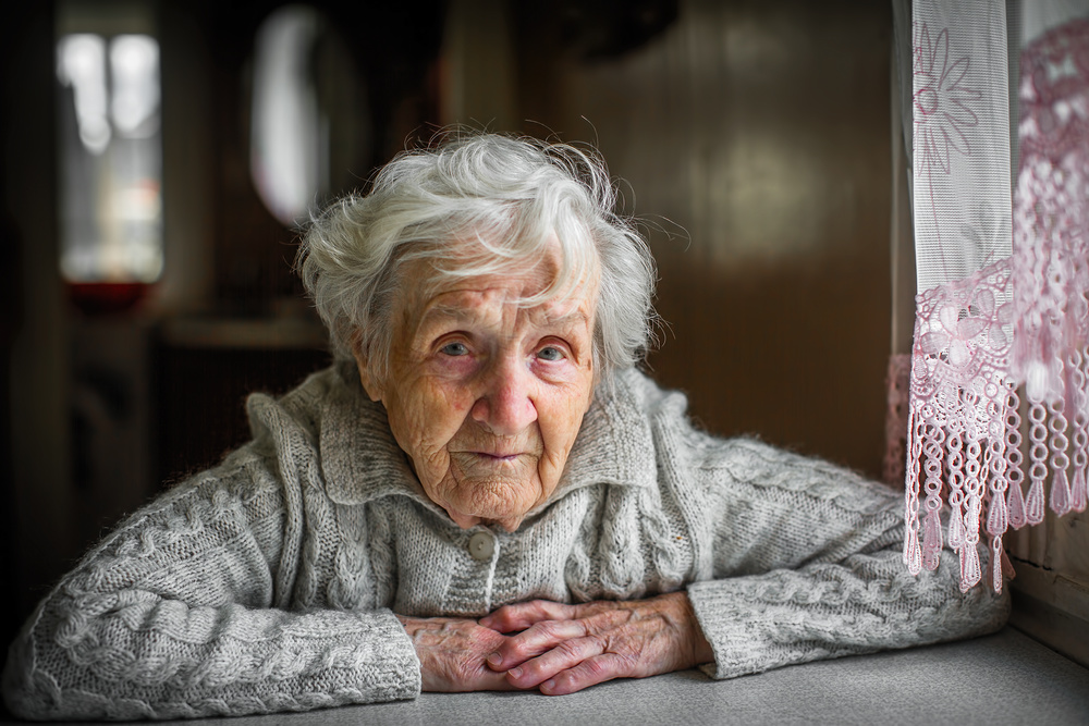 Gray-haired elderly woman sitting near the window. / ©De Visu - stock.adobe.com, Dmitry Berkut Gray-haired elderly woman sitting near the window.